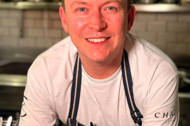 Portrait of Chef Tom de Keyser wearing a striped Hand & Flowers apron, smiling in a kitchen ahead of the London 4-hand dinner collaboration.