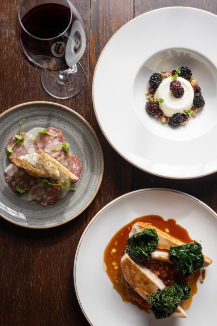 A three-course Autumn Table meal featuring a cured meat and crostini starter, a sliced roasted poultry main course with dark leafy greens, and a blackberry panna cotta dessert, served with a glass of red wine.
