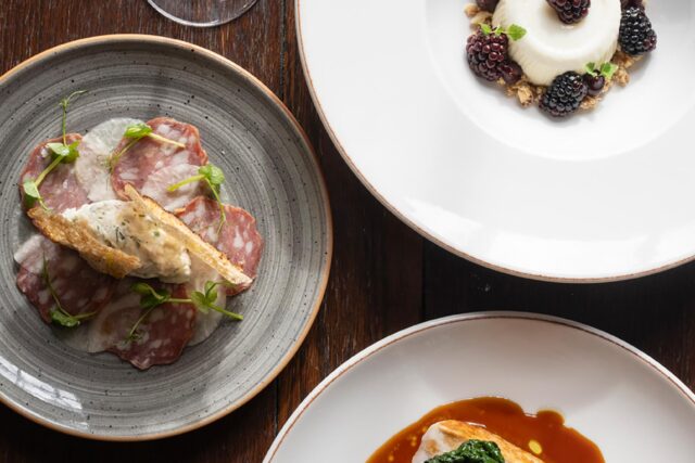 A three-course Autumn Table meal featuring a cured meat and crostini starter, a sliced roasted poultry main course with dark leafy greens, and a blackberry panna cotta dessert, served with a glass of red wine.