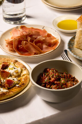 Autumn Table starters at Galvin Bistrot & Bar, showcasing a fig and cheese flatbread with pine nuts, a bowl of baby squid in tomato sauce, a plate of thinly sliced cured ham, and fresh bread with olive oil.