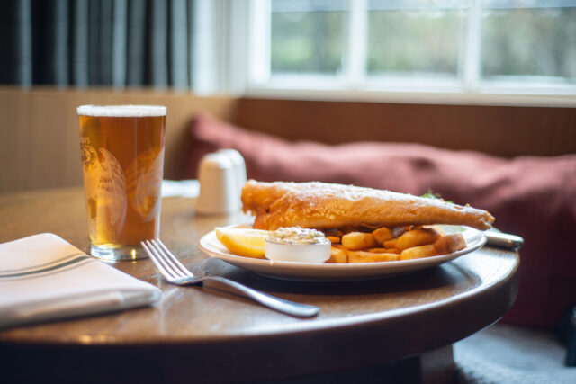 A portion of battered fish and chips accompanied by a pint of beer, resting on a round wooden pub table by a window.