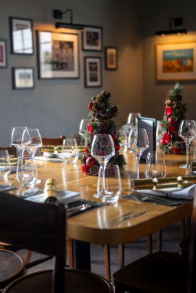 A warm and festive dining room table set for Christmas at Galvin Green Man, featuring miniature decorated Christmas trees, gold crackers, and polished wine glasses.