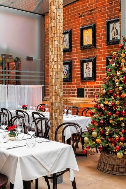 The glass-roofed dining room at Galvin Spitalfields prepared for a festive event, featuring set tables with white linens, dark wood chairs, exposed brick walls, and a fully decorated Christmas tree.