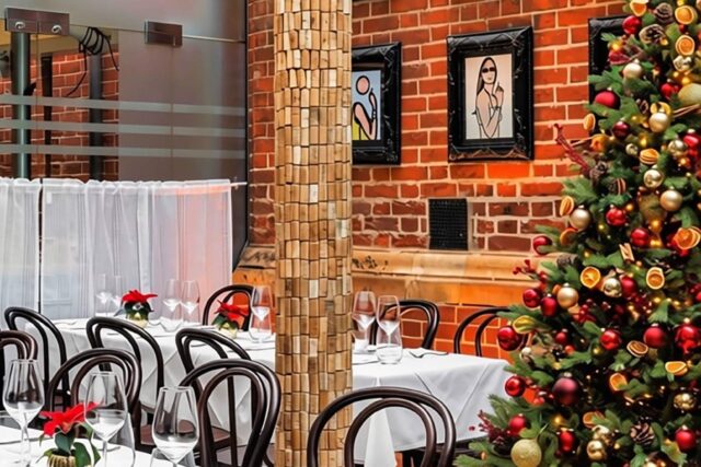 The glass-roofed dining room at Galvin Spitalfields prepared for a festive event, featuring set tables with white linens, dark wood chairs, exposed brick walls, and a fully decorated Christmas tree.