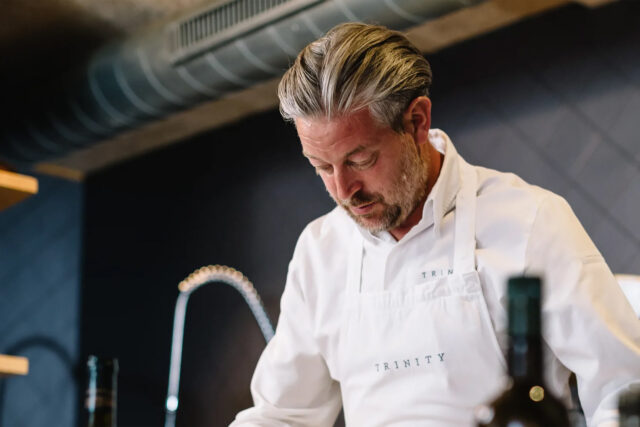 Chef Adam Byatt wearing a white Trinity apron preparing food in a kitchen setting.